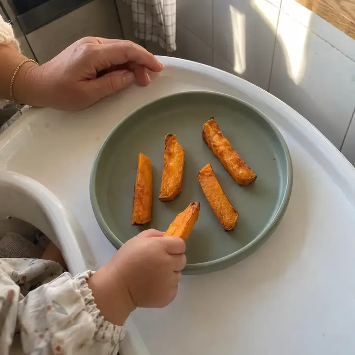 Bastones de boniato asado en un plato de silicona sobre la bandeja de la trona; la mano del bebé coge uno y la de su madre sujeta la bandeja