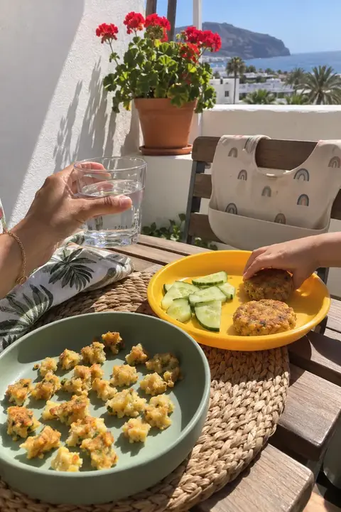 Mesa en un balcón soleado con hamburguesitas de garbanzos: troceadas en el plato del bebé y enteras en el plato de su hermano mayor