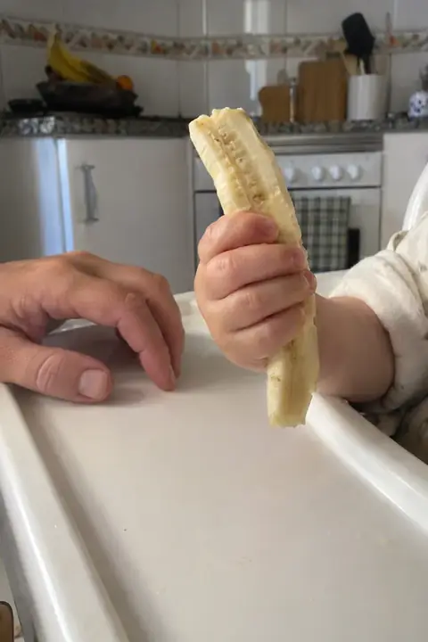 Mano de bebé cogiendo una tira de plátano sobre la bandeja de la trona