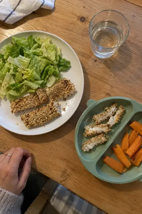 Mesa familiar vista desde arriba: el plato del adulto con palitos de merluza y ensalada y, al lado, el plato de silicona del bebé con la misma merluza en tiras